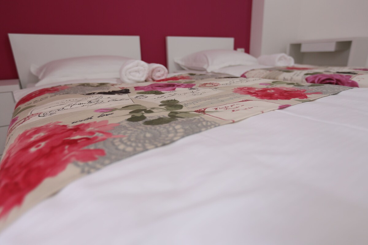 A neatly made bed with a floral quilt, white pillows, and a pink wall in the background.