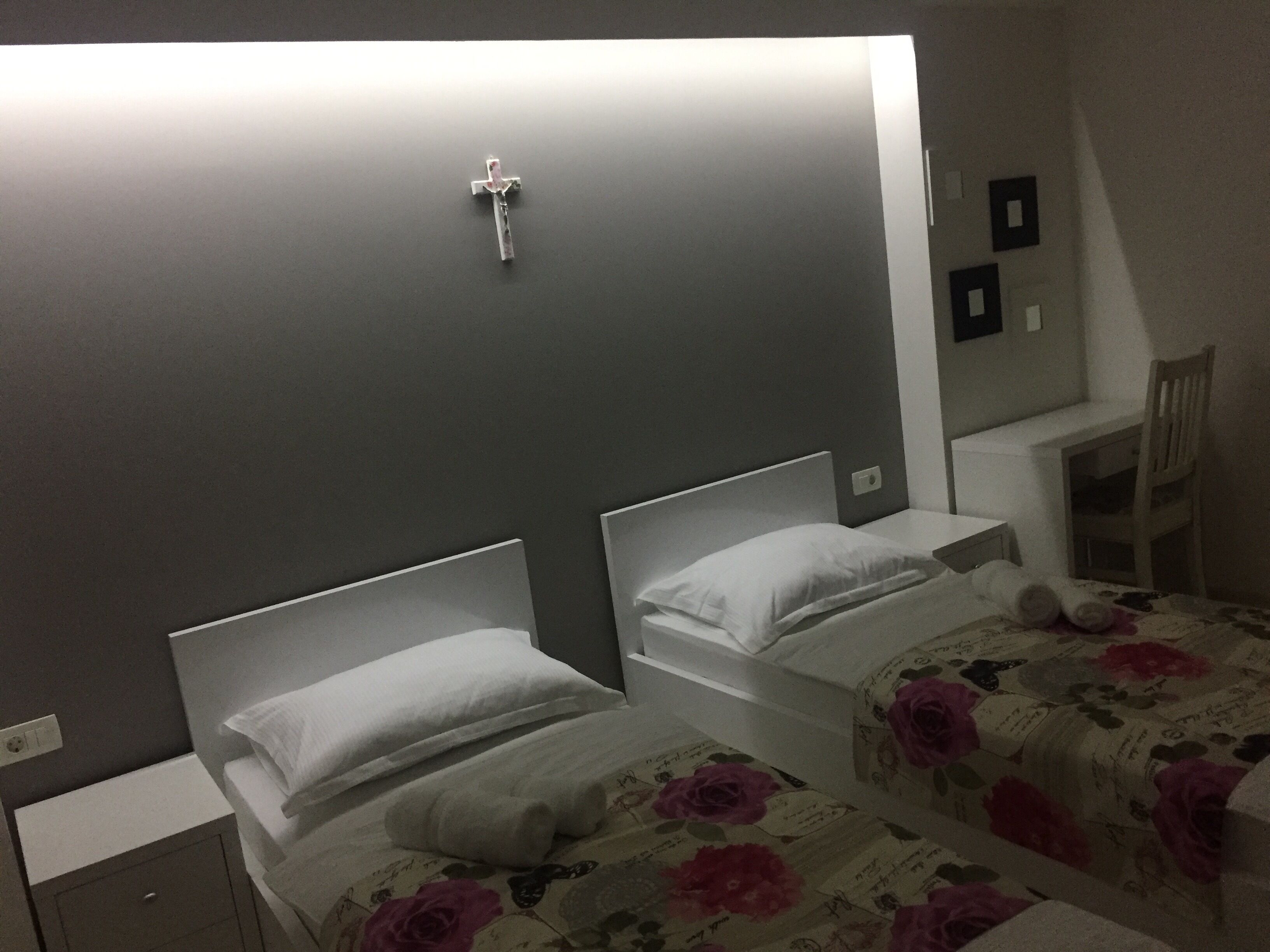 A hotel room with two beds, a small desk, a chair, and a cross on the wall.