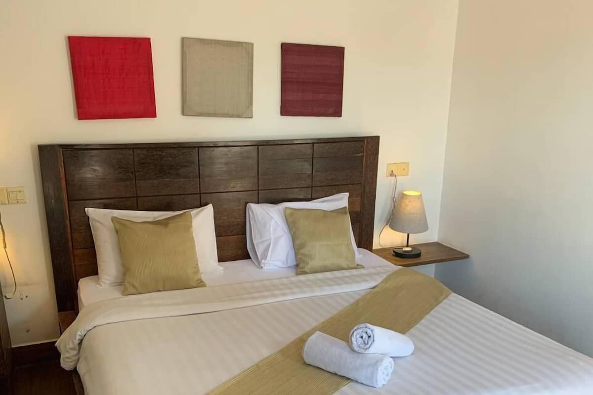 Una cama bien hecha con cabecera de madera, ropa de cama blanca y beige, y una toalla enrollada sobre la cama. Hay tres obras de arte colgadas en la pared encima de la cama.