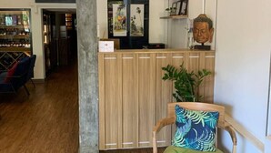 A reception area with a wooden reception desk, a chair with a green cushion and a blue patterned pillow, a small round table with a potted plant, and a store named 'MONSIEUR' in the background. - Suite Home Boutique Hotel (Phnom Penh)