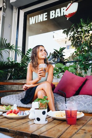 A person sitting on a bench outside a wine bar, holding a drink, with a table in front of them containing food and a drink. - Suite Home Boutique Hotel (Phnom Penh)