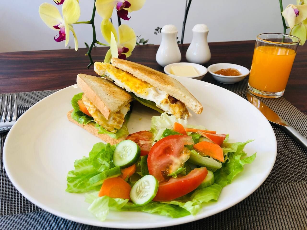 Un plato con un sándwich y una ensalada, un vaso de jugo de naranja y aderezos sobre una mesa.
