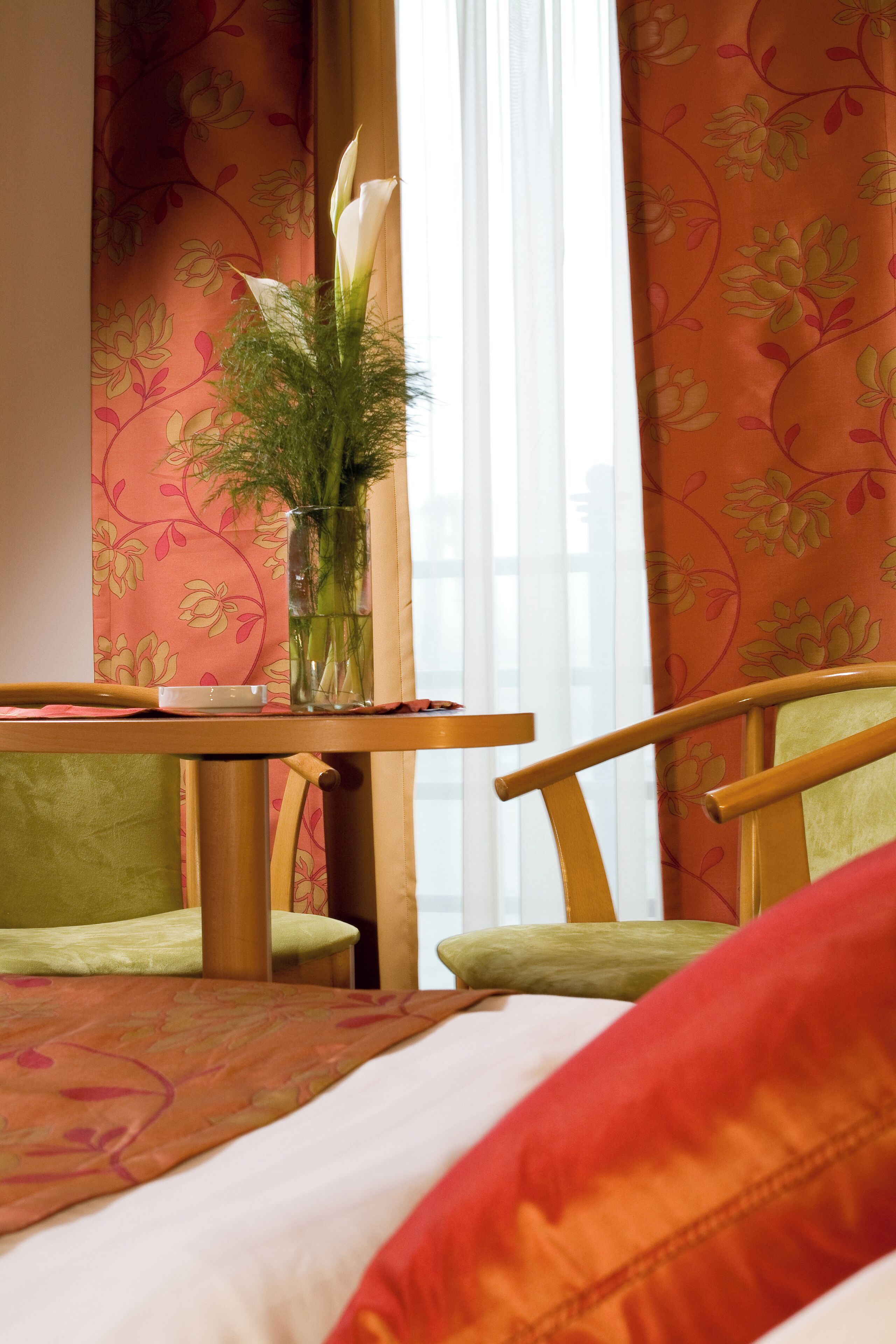A hotel room with a wooden table, two green chairs, a vase with a plant, and a floral-patterned wall.
