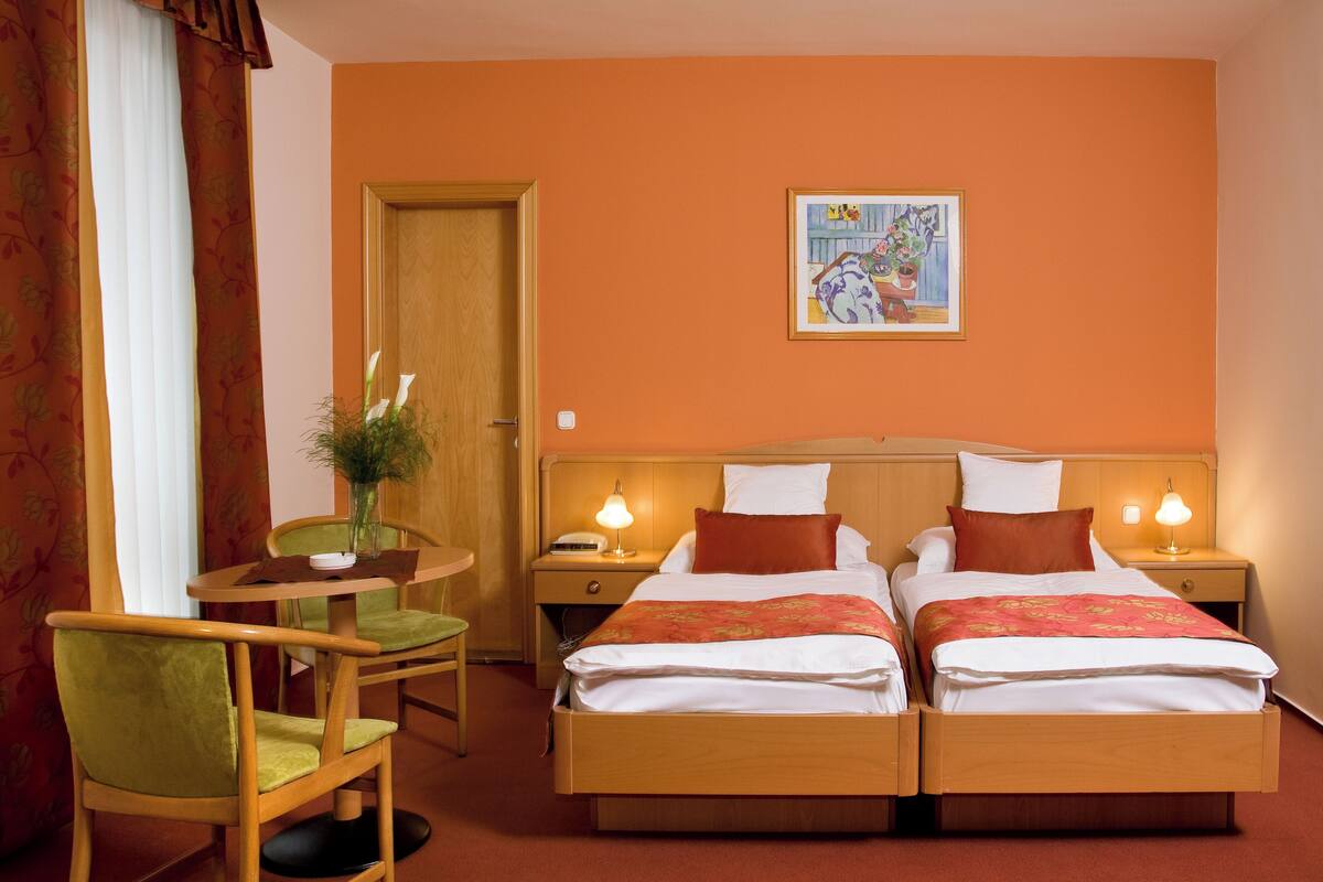 Habitación de hotel con dos camas, cabecero de madera, una mesita redonda pequeña con una silla, una mesita de noche con lámpara y un cuadro en la pared.