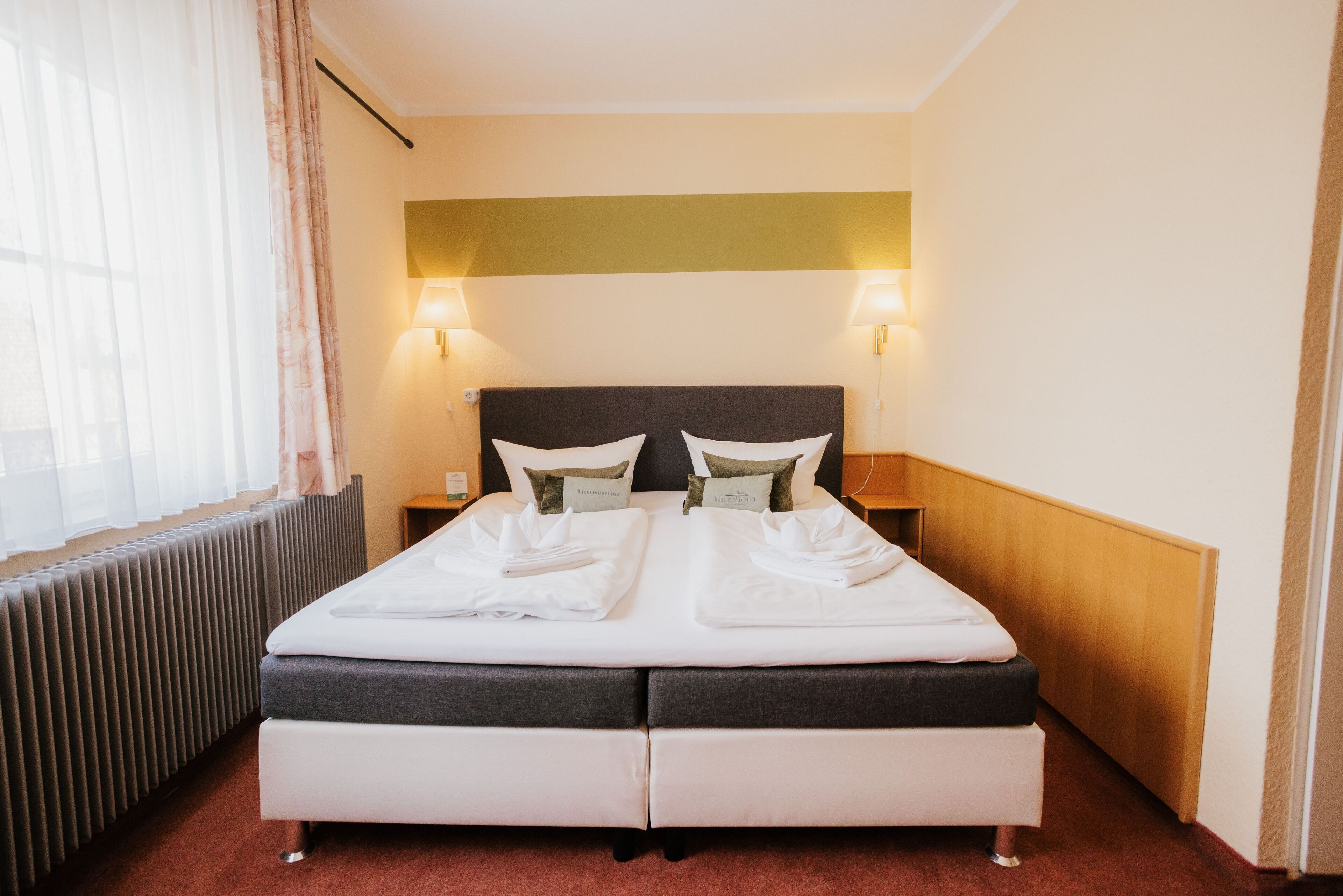 Een hotelkamer met een tweepersoonsbed, twee bedlampjes, een radiator en een raam met gordijnen.
