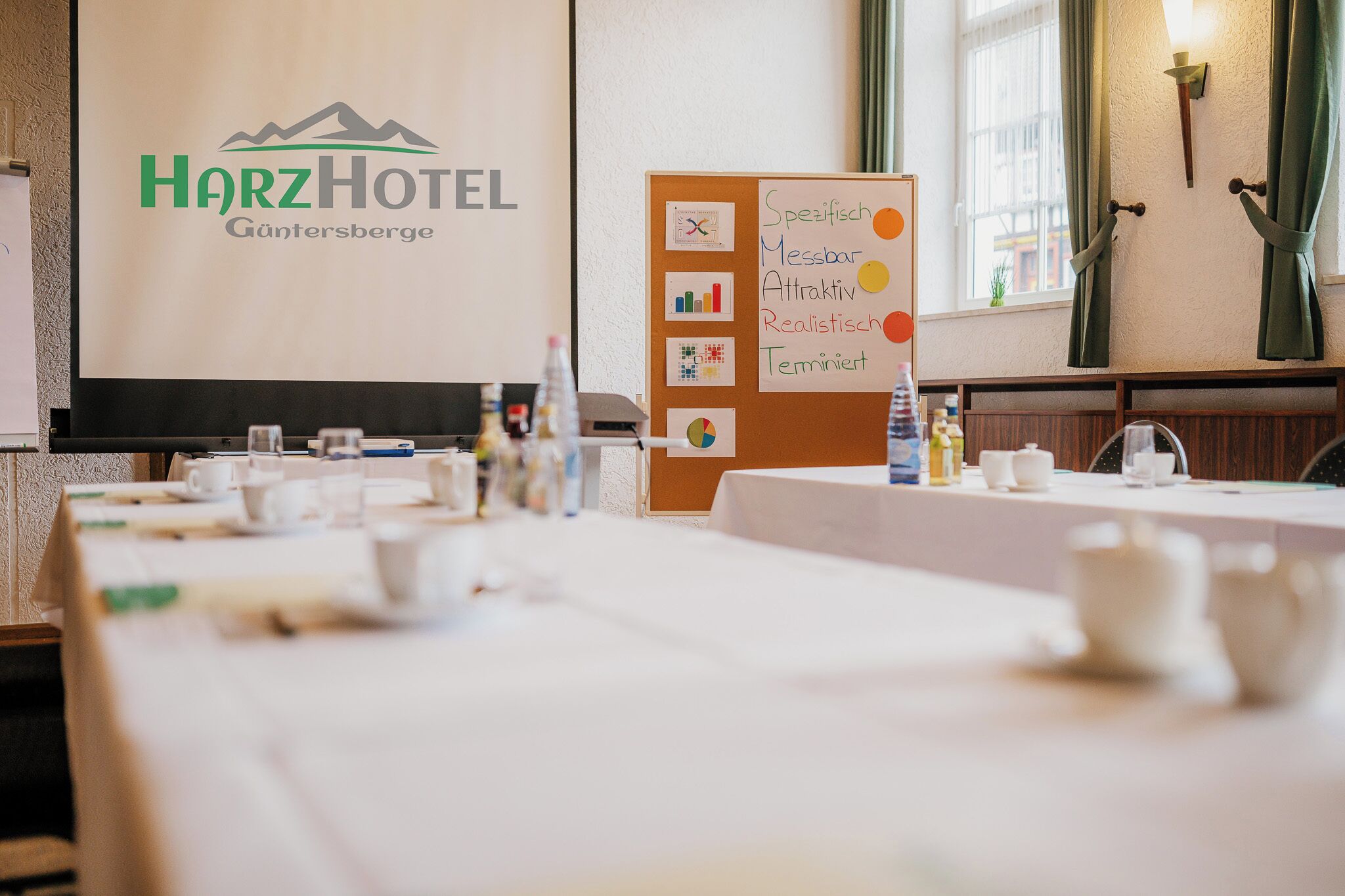 A conference room at HarzHotel in Gunnersberg with tables set for a meeting, a presentation screen, and a bulletin board with various charts.