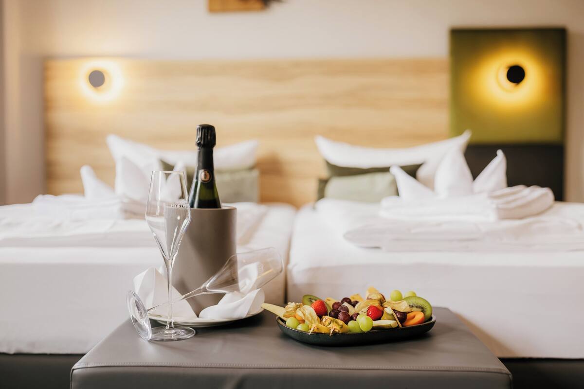 A hotel room with a bed, a bedside table with a bottle of champagne, a glass, and a fruit bowl.