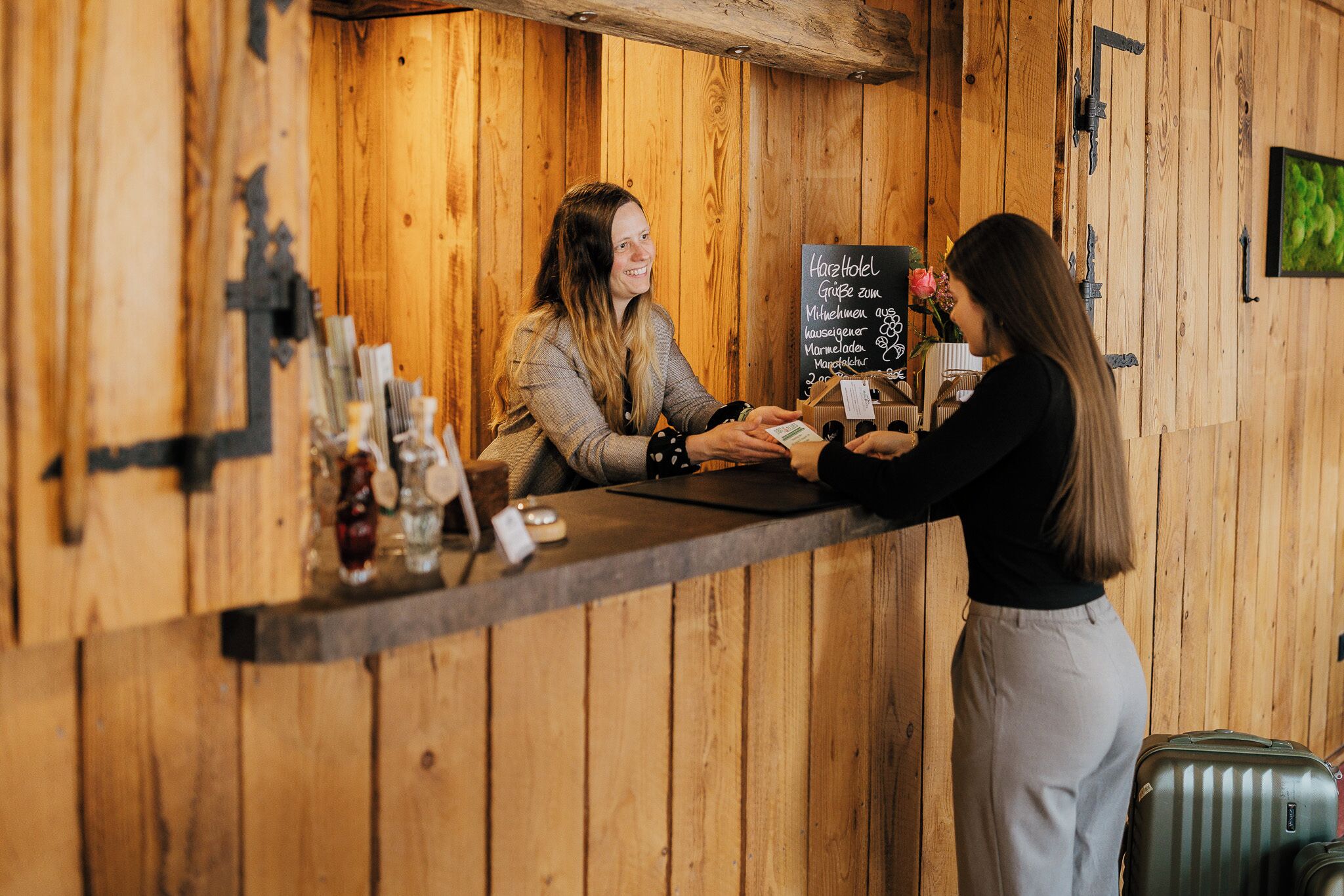 A wooden reception desk with a chalkboard menu, a bar area with bottles, and a suitcase on the floor.