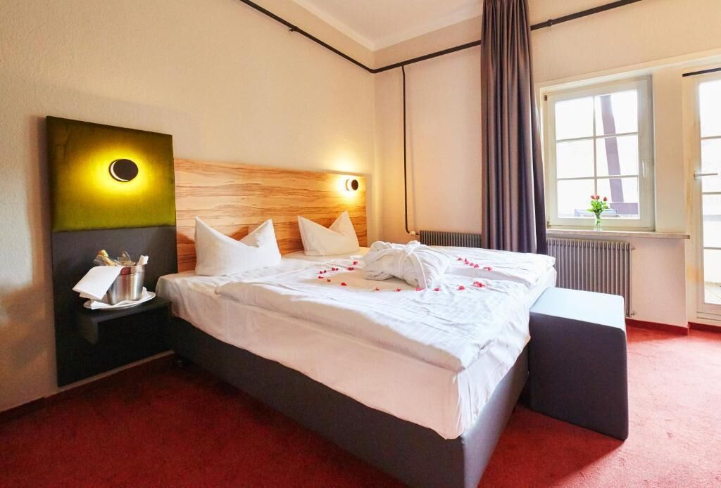 Una habitación de hotel con una cama grande, una mesita de noche con lámpara, una ventana con cortinas y una obra de arte colgada en la pared.