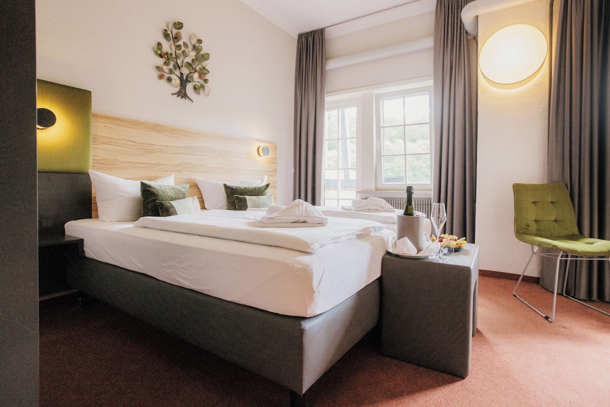 A modern hotel room with a large bed, a bedside table with a lamp, a chair, and a view of the outdoors through a window.