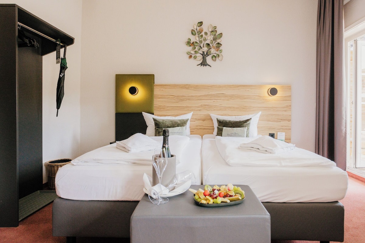 A hotel room with two beds, a bedside table with a fruit plate, a bottle of champagne, and a wall-mounted artwork.