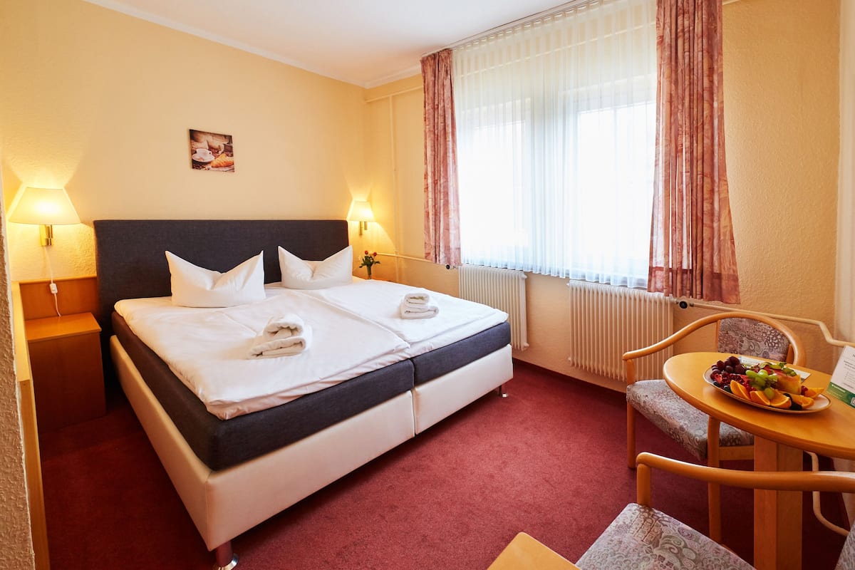 A hotel room with a bed, two bedside lamps, a table with fruit, a chair, and a window with curtains.