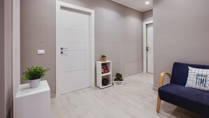 A hallway with a white door, a small white shelf with books and plants, a blue sofa with a pillow, and a wooden bench. - Oasis (Aci Castello)