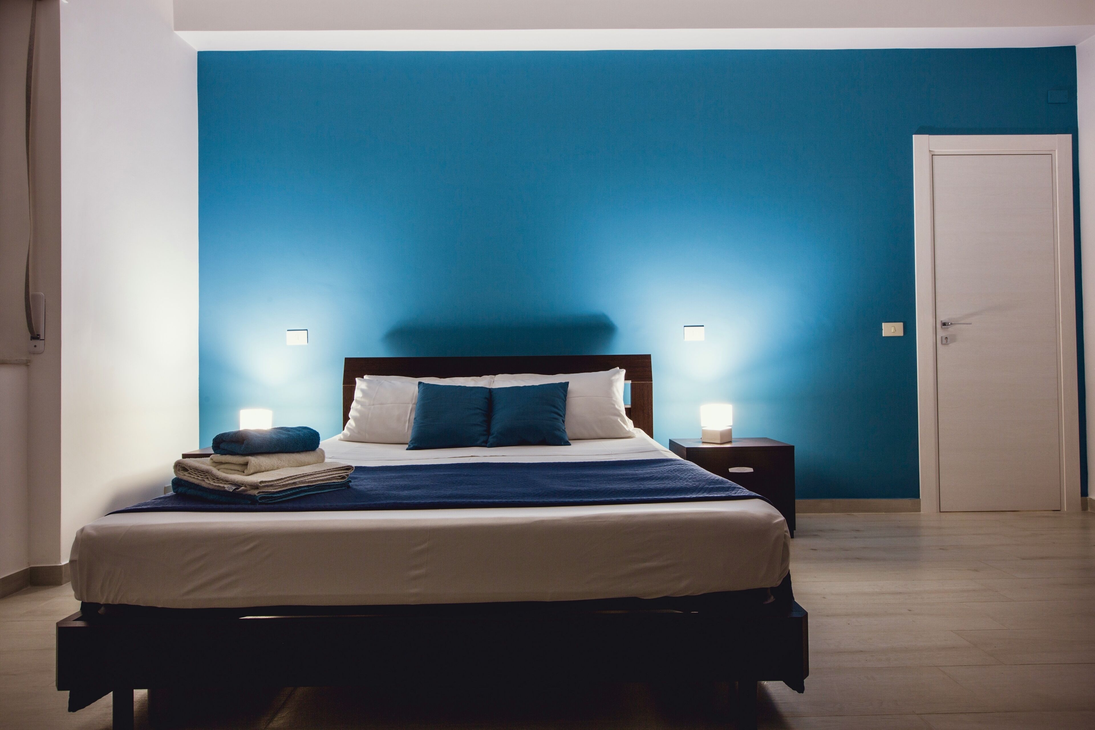 Una cama bien hecha con cabecera azul, ropa de cama blanca y almohada azul. Hay dos lámparas de noche a cada lado de la cama.