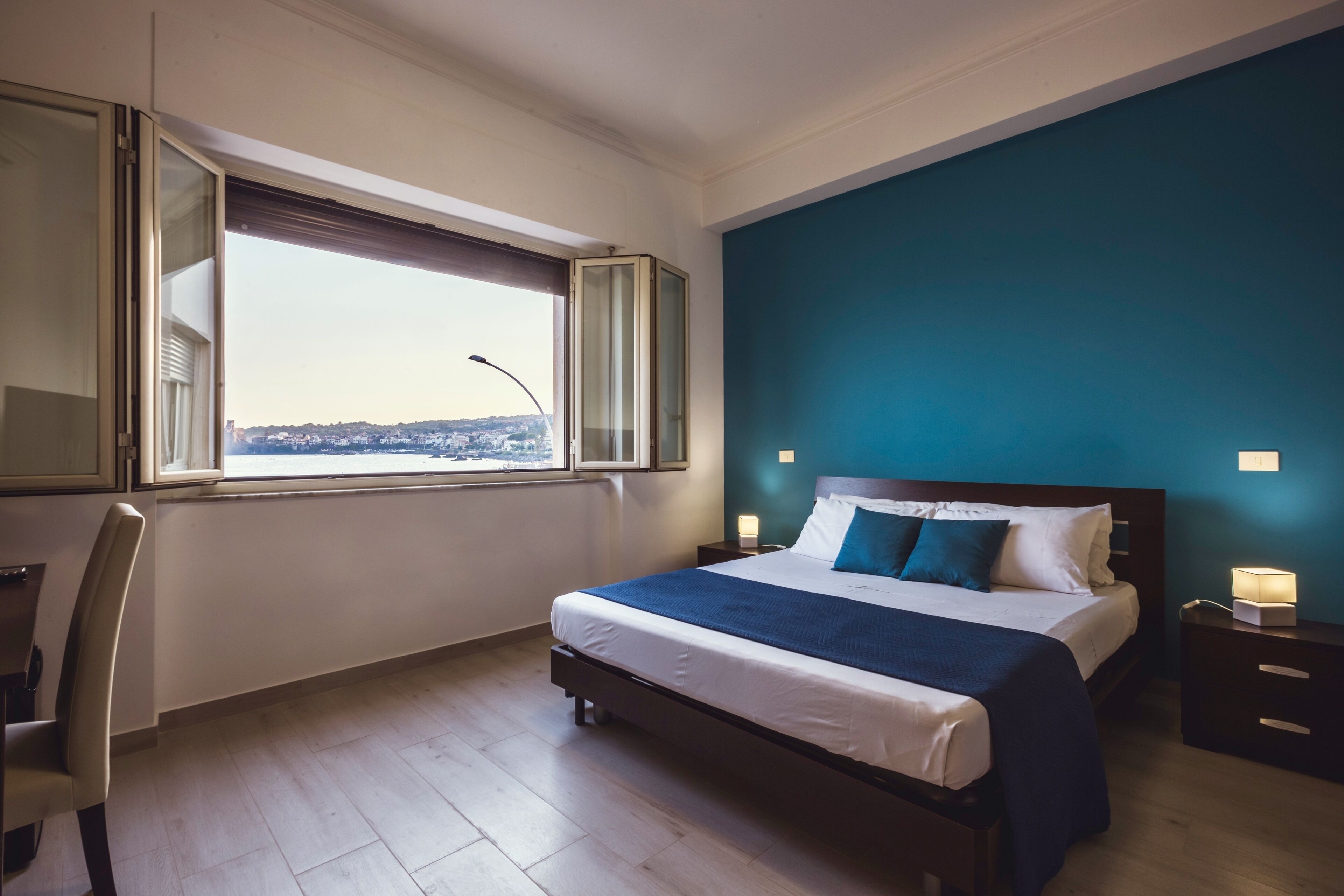 Ein Schlafzimmer mit Bett, Stuhl, Nachttisch, Lampe und einem Fenster mit Blick auf eine Stadtlandschaft.