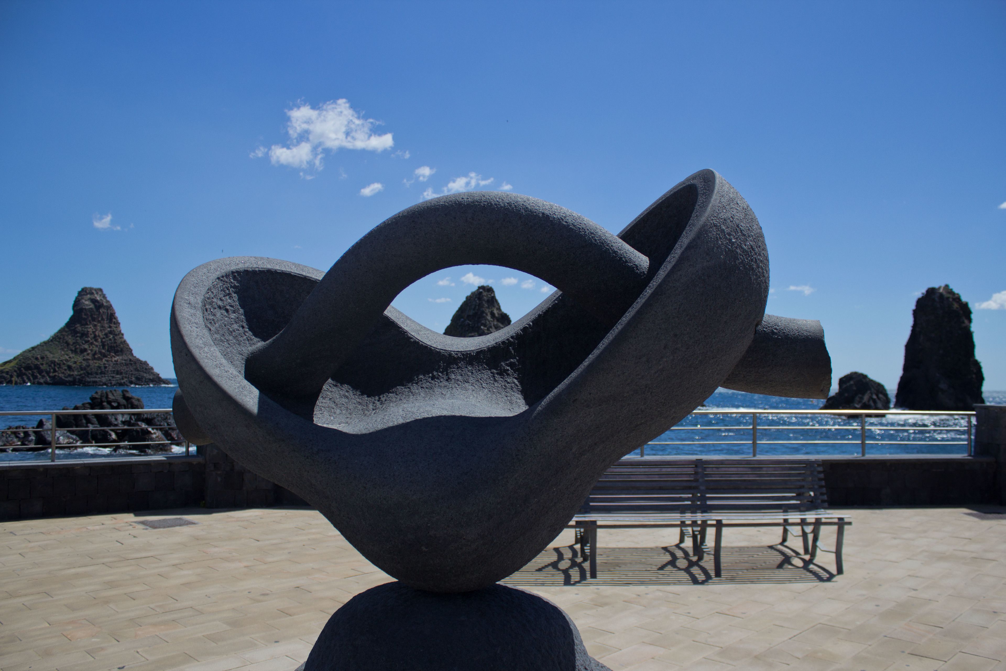 Una escultura en un entorno exterior con un banco y vistas al mar y formaciones rocosas.