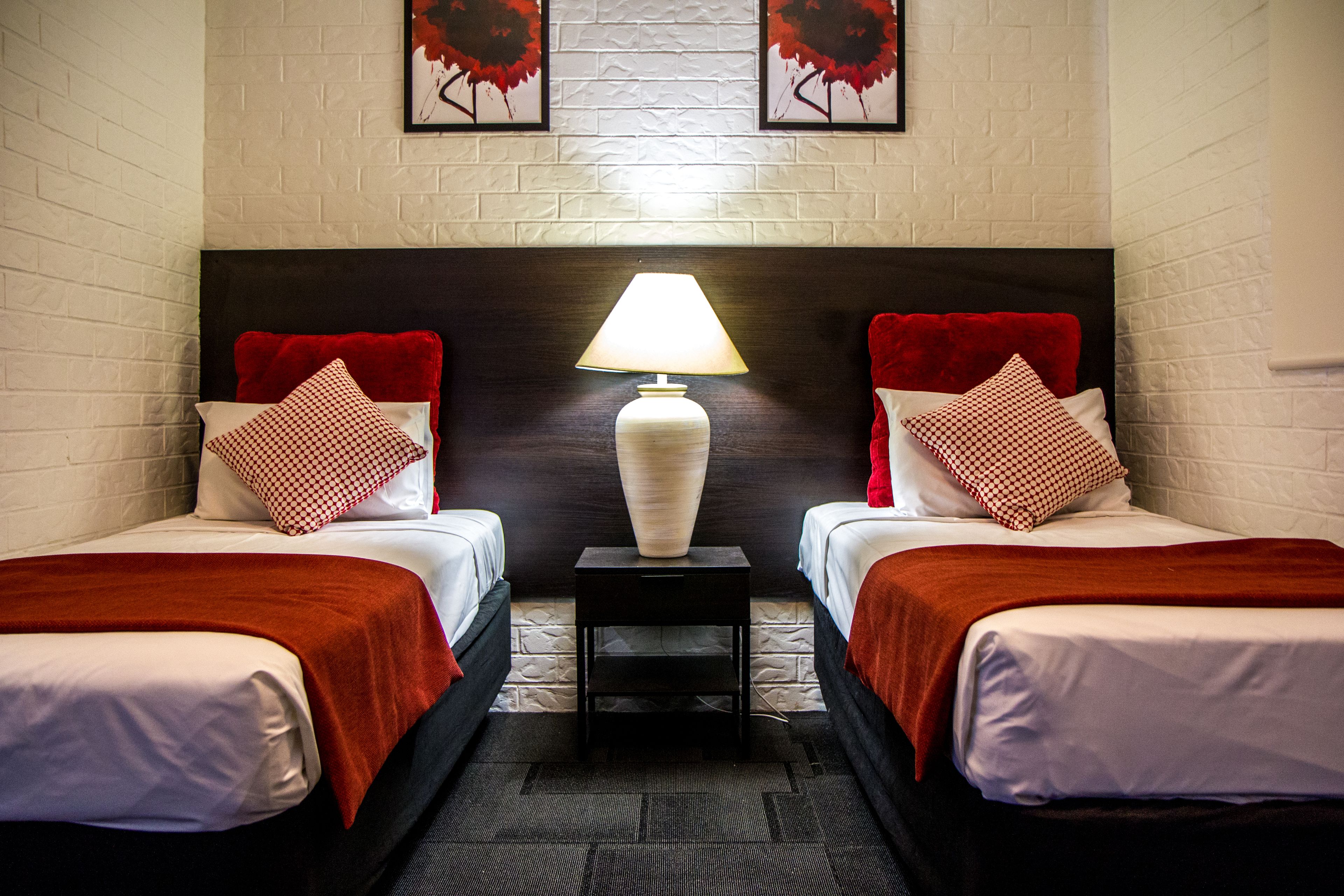 A hotel room with two beds, each with a red and white checkered pillow, a white vase lamp, and a brick-patterned wall.