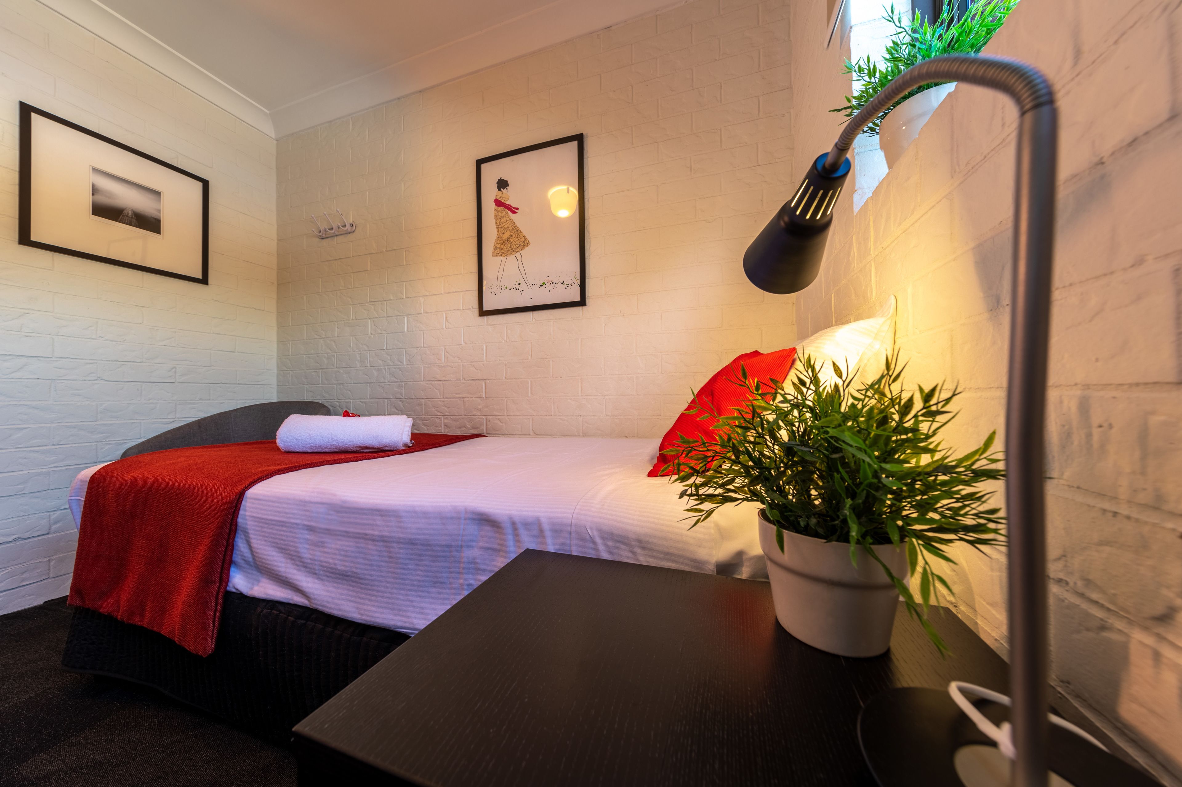 Un dormitorio ordenado con cama, mesita de noche, lámpara y planta en maceta.