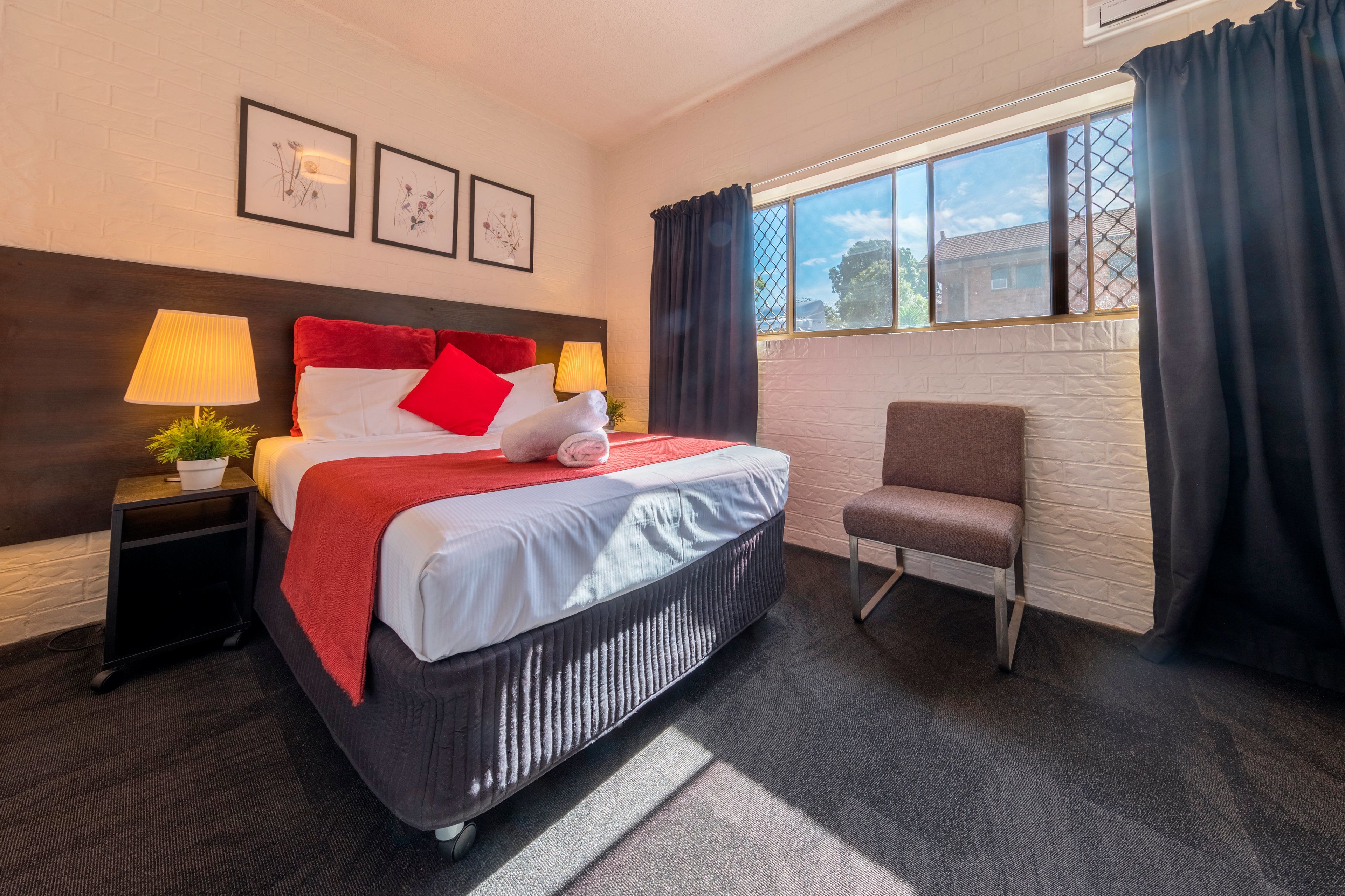 A hotel room with a bed, a chair, a nightstand with a lamp, and a window with curtains.