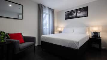 Una habitación de hotel moderna con una cama grande, dos lámparas de noche, un sofá gris con un cojín rojo, una mesita con una planta y un cuadro en la pared.