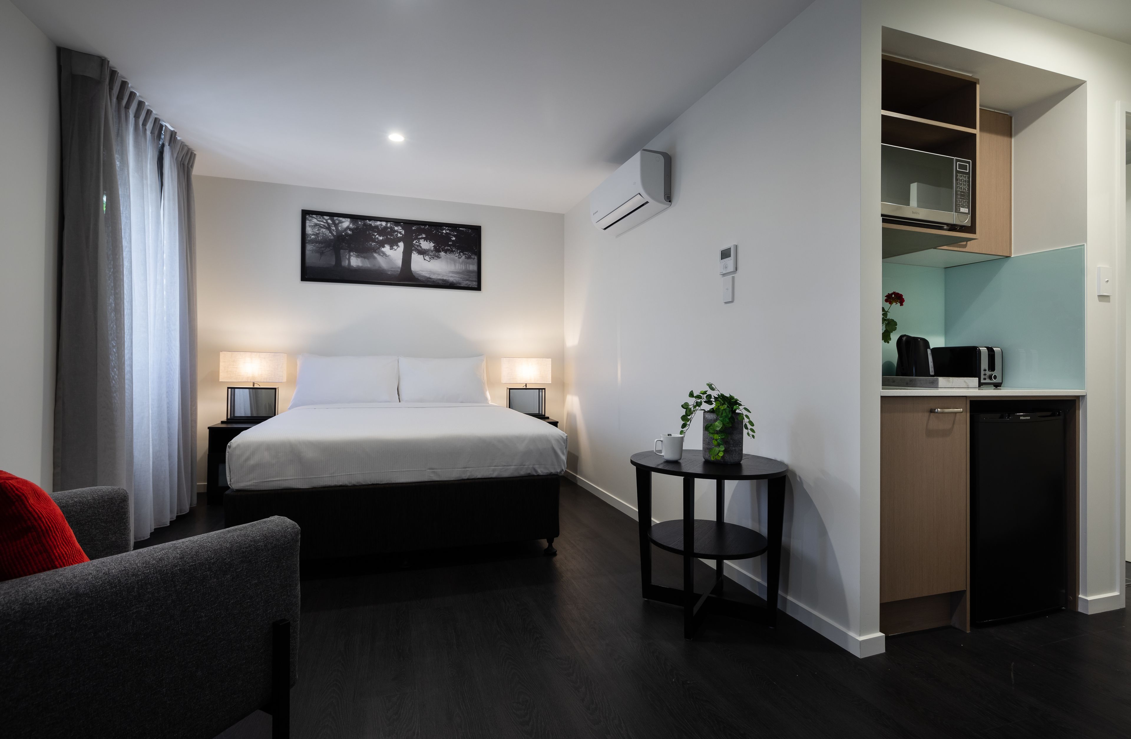 A hotel room with a bed, bedside lamps, a small table with a plant, a desk with a microwave, and a compact kitchenette.
