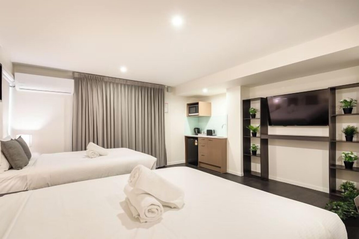 Une chambre d’hôtel avec deux lits, une télévision, une kitchenette et une étagère avec des plantes.