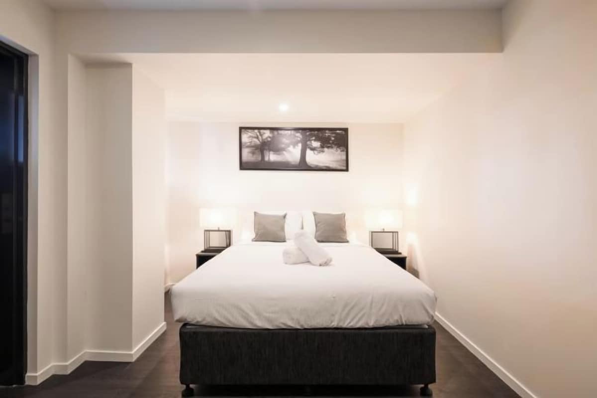 A hotel room with a large bed, two bedside tables with lamps, a framed picture on the wall, and a dark-colored bench at the foot of the bed.