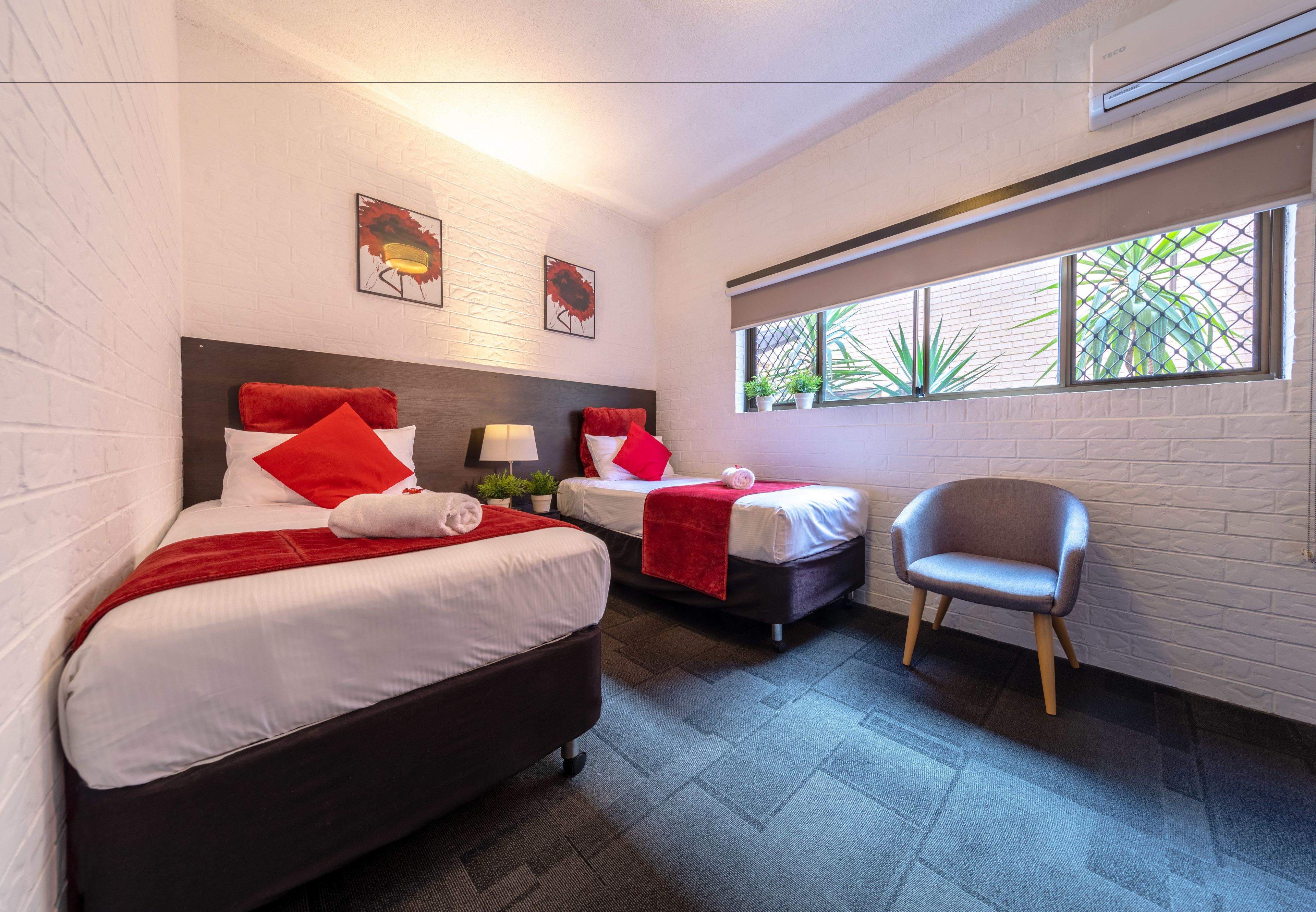 A hotel room with two beds, a chair, and a window with plants.