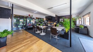 A modern office space with wooden flooring, a central dining area with tables and chairs, and potted plants.