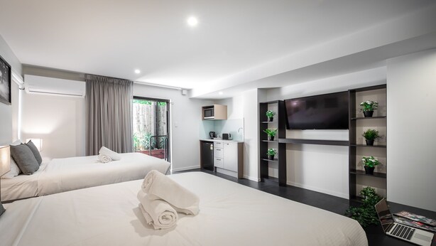 A modern hotel room with two beds, a TV, a desk, and a balcony with plants.