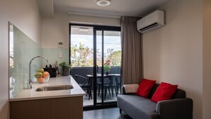 A modern kitchen with a sink, a dining area with a table and chairs, a grey sofa with red cushions, and a sliding glass door leading to a balcony with a view of trees and a building.