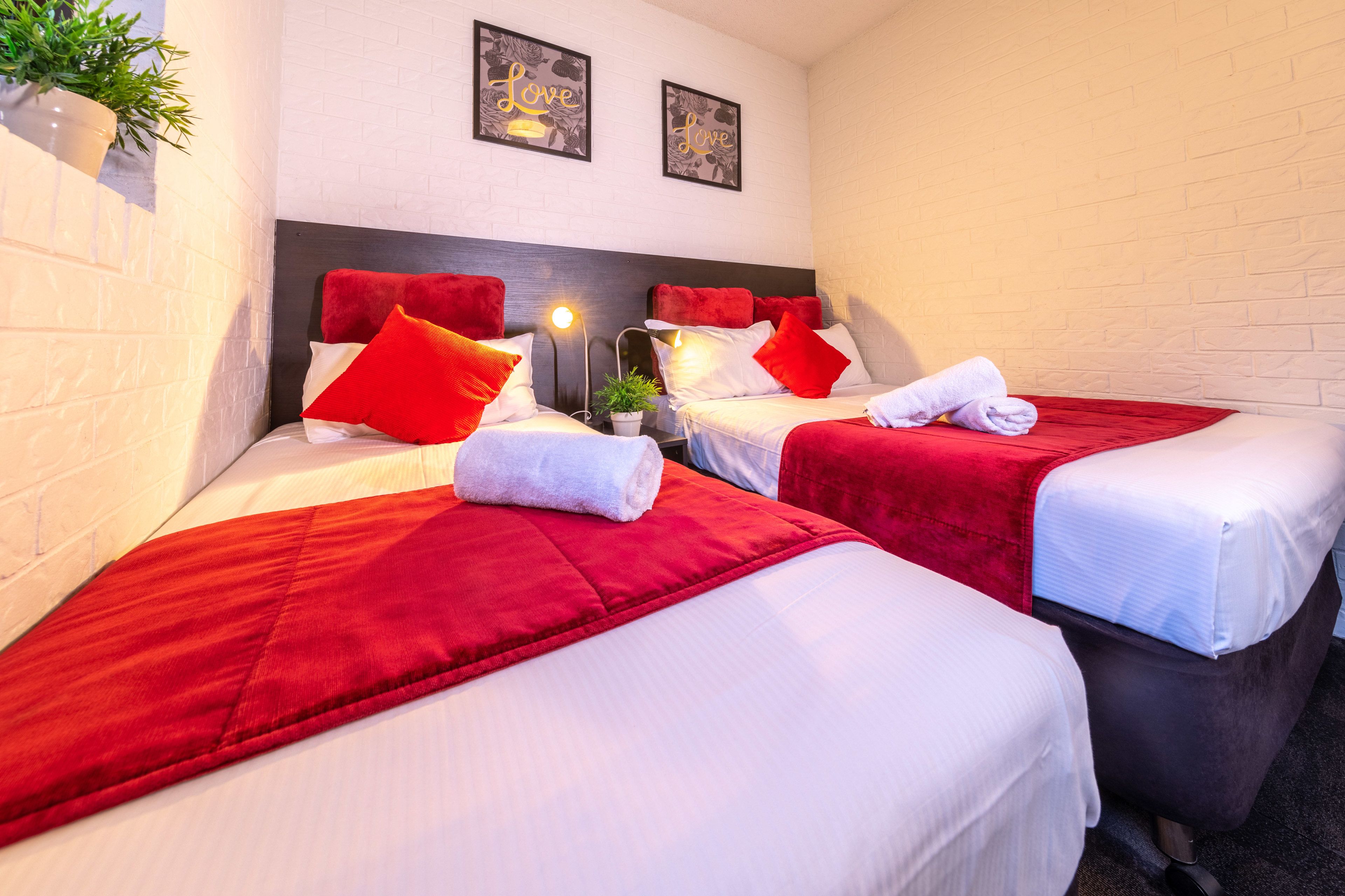 Une chambre d’hôtel avec deux lits, chacun agrémenté de draps rouges et d’oreillers blancs. Deux tableaux encadrés ornent le mur, une plante en pot se trouve à gauche et une lampe à droite.