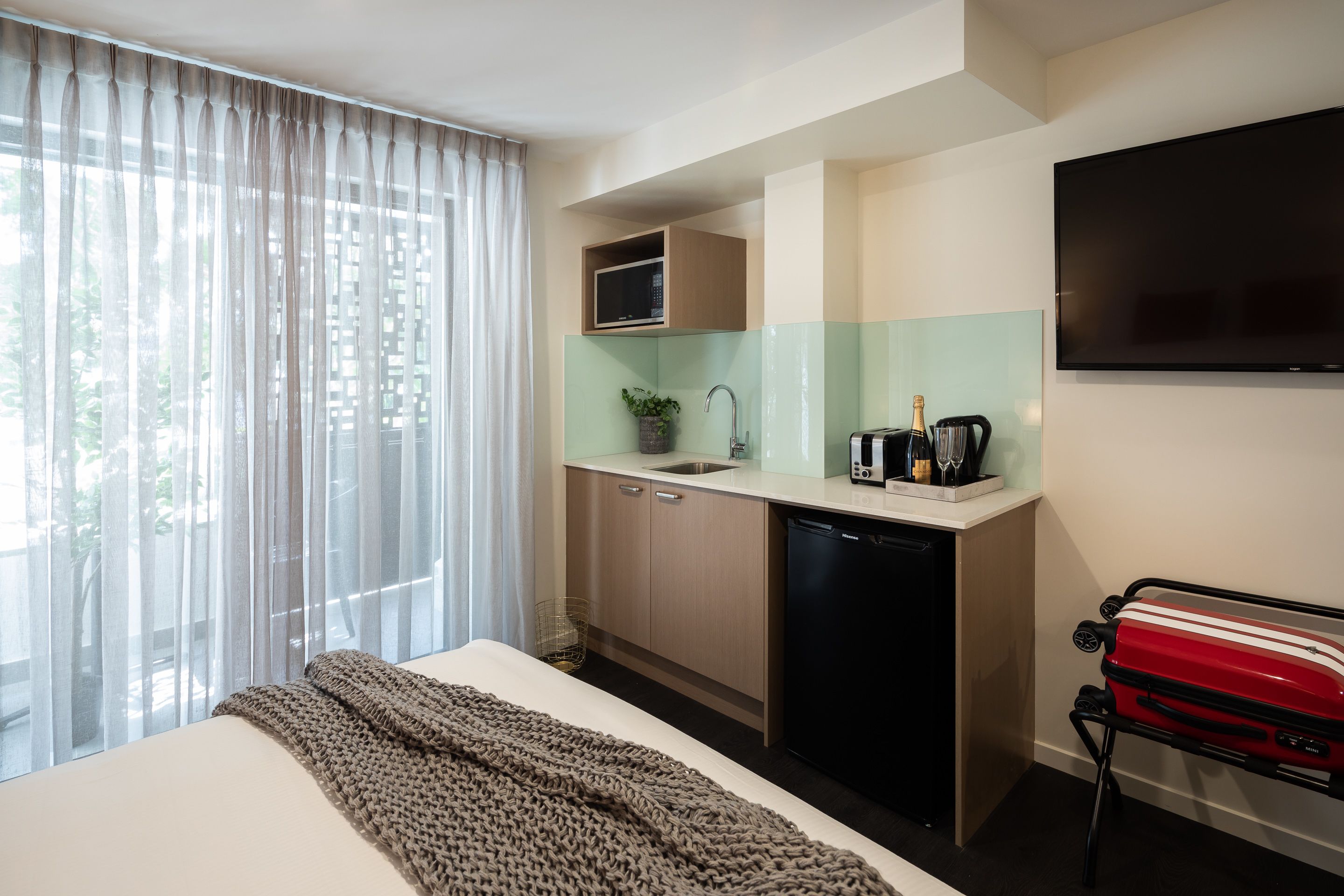Een kleine hotelkamer met een bed, een kitchenette, een televisie en een koffer.