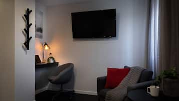 Una sala de estar moderna con un televisor de pantalla plana montado en la pared, un sillón gris, un sofá con un cojín rojo, una mesita con una taza blanca y un escritorio con una computadora portátil y una lámpara.