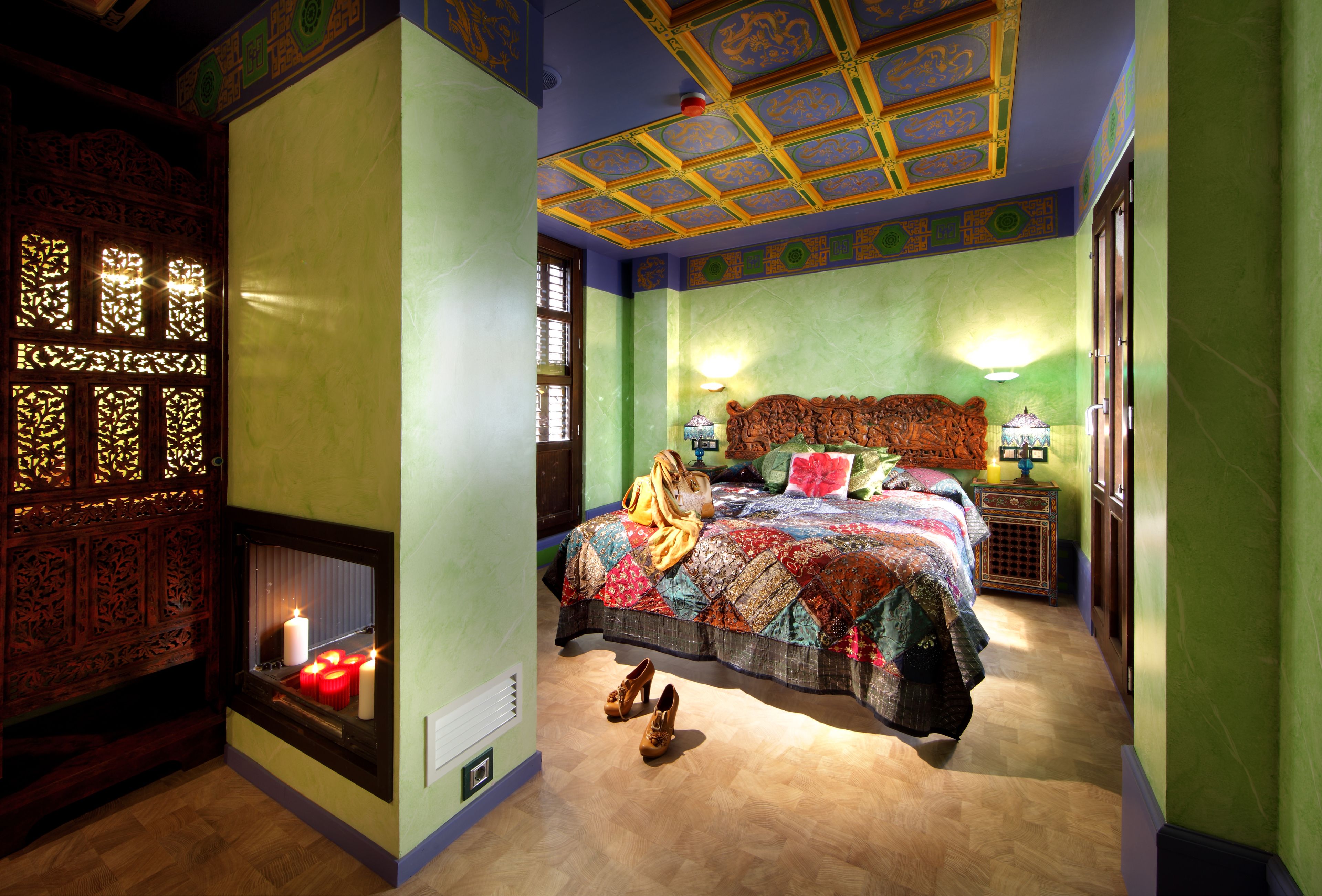Una habitación con una cama, una ventana con persianas decorativas, un calentador de pared y una pequeña mesa con lámpara.