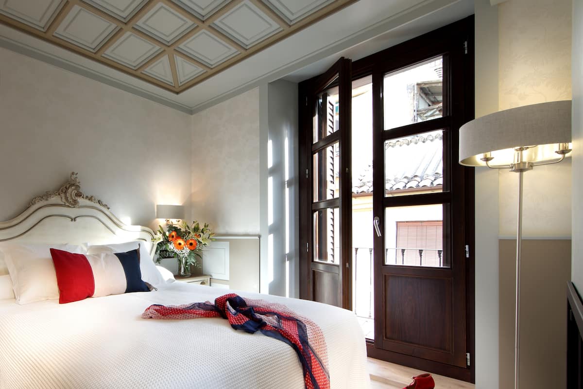 A neatly made bed with pillows, a bedside table with a lamp, and a view of the outside through a large window.