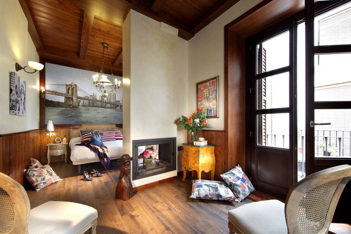 A cozy room with a bed, a fireplace, a wooden floor, and a large window.