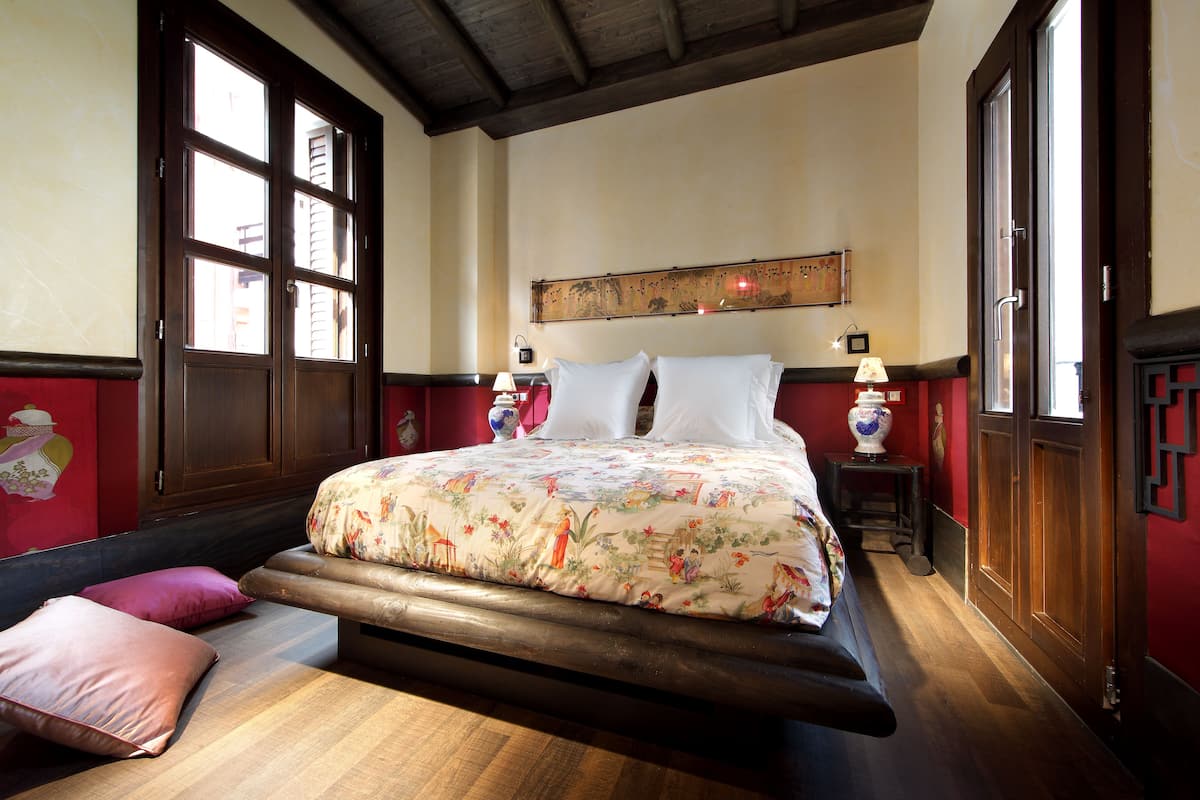 A bedroom with a large bed, wooden headboard, and a floral bedspread. There are two bedside tables with lamps and a decorative vase.
