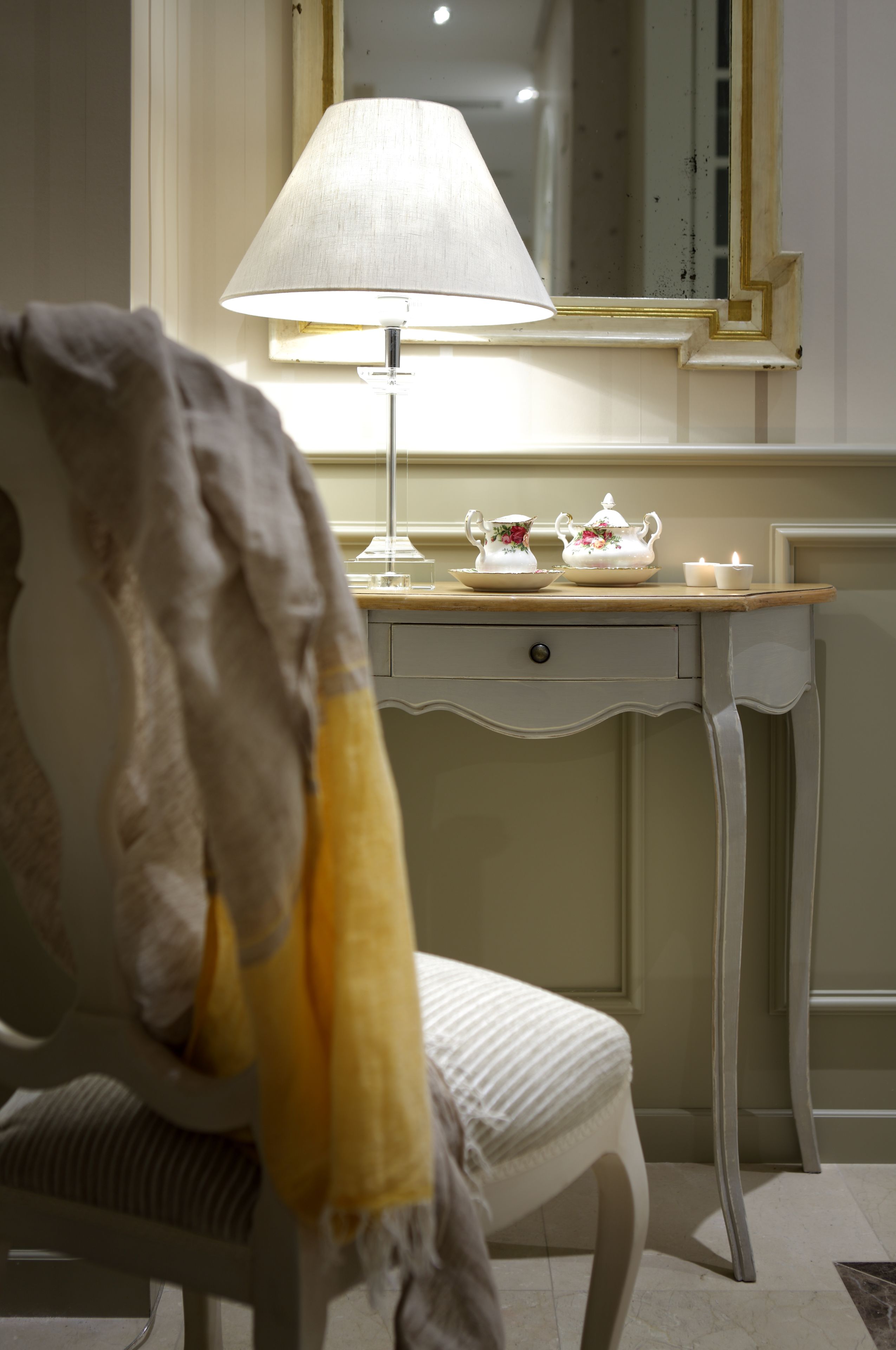 Una habitación con una silla blanca, una mesa con una lámpara y un espejo.