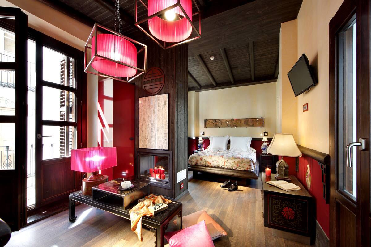 A hotel room with a bed, bedside table, lamp, and a view of the city through large windows.