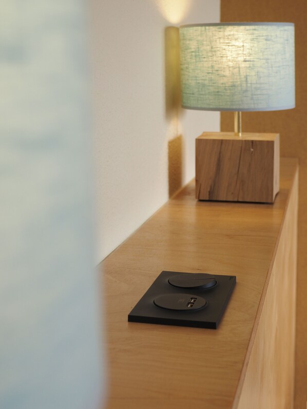A wooden bedside table with a modern lamp and a small electronic device.