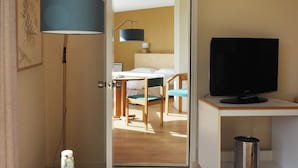 A hotel room with a bed, a television, a table with a bottle and glasses, a lamp, and a door leading to a dining area. - Belvédère Hôtel (Brest)