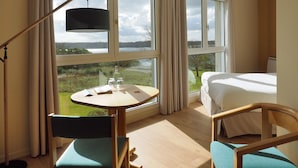A hotel room with a bed, a desk with a chair, a table with a chair, and large windows offering a view of the outdoors. - Belvédère Hôtel (Brest)