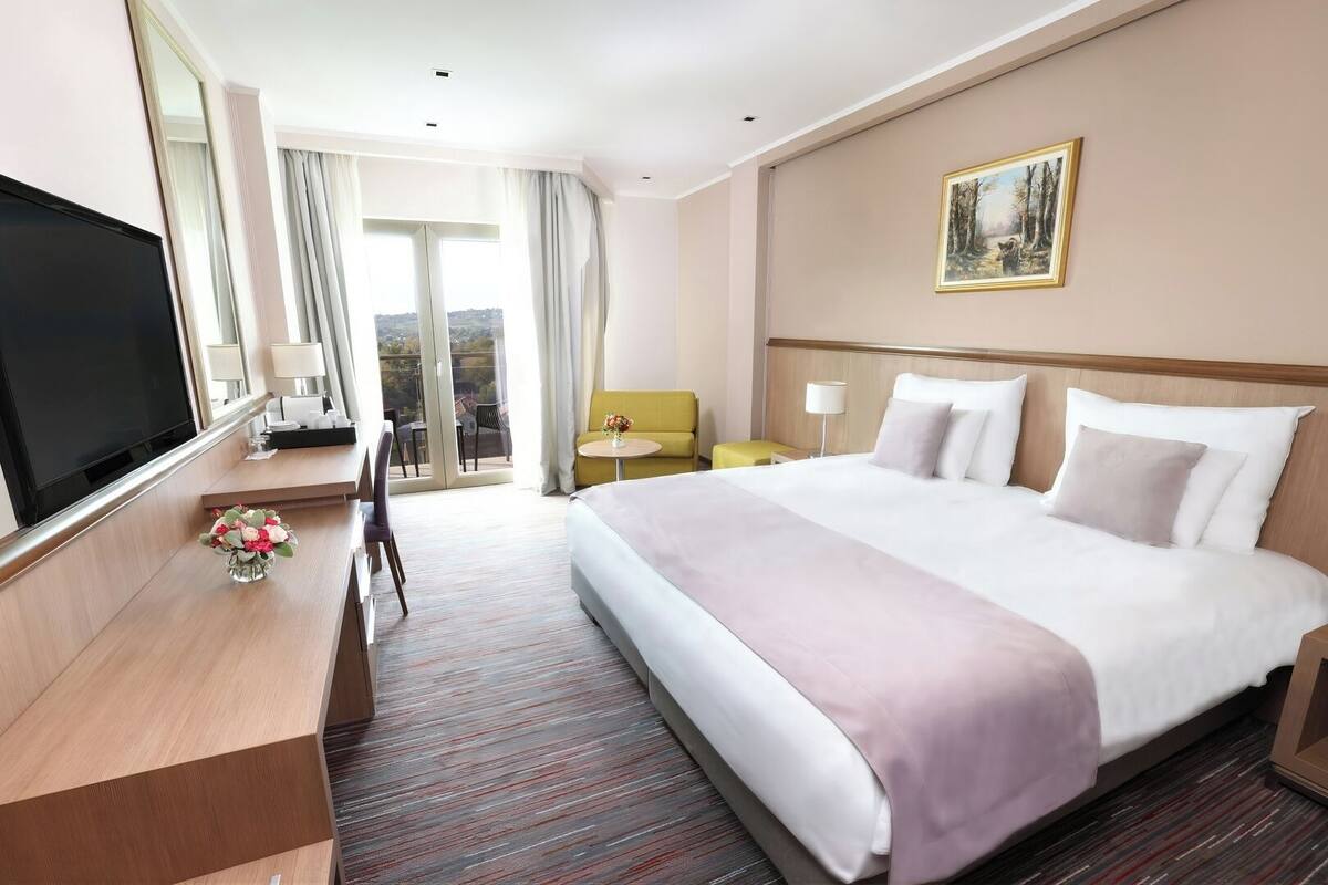 Ein Hotelzimmer mit einem großen Bett, einem Flachbildfernseher, einem Schreibtisch mit Stuhl, einem kleinen Tisch mit Lampe und Blick ins Freie durch eine Schiebetür.