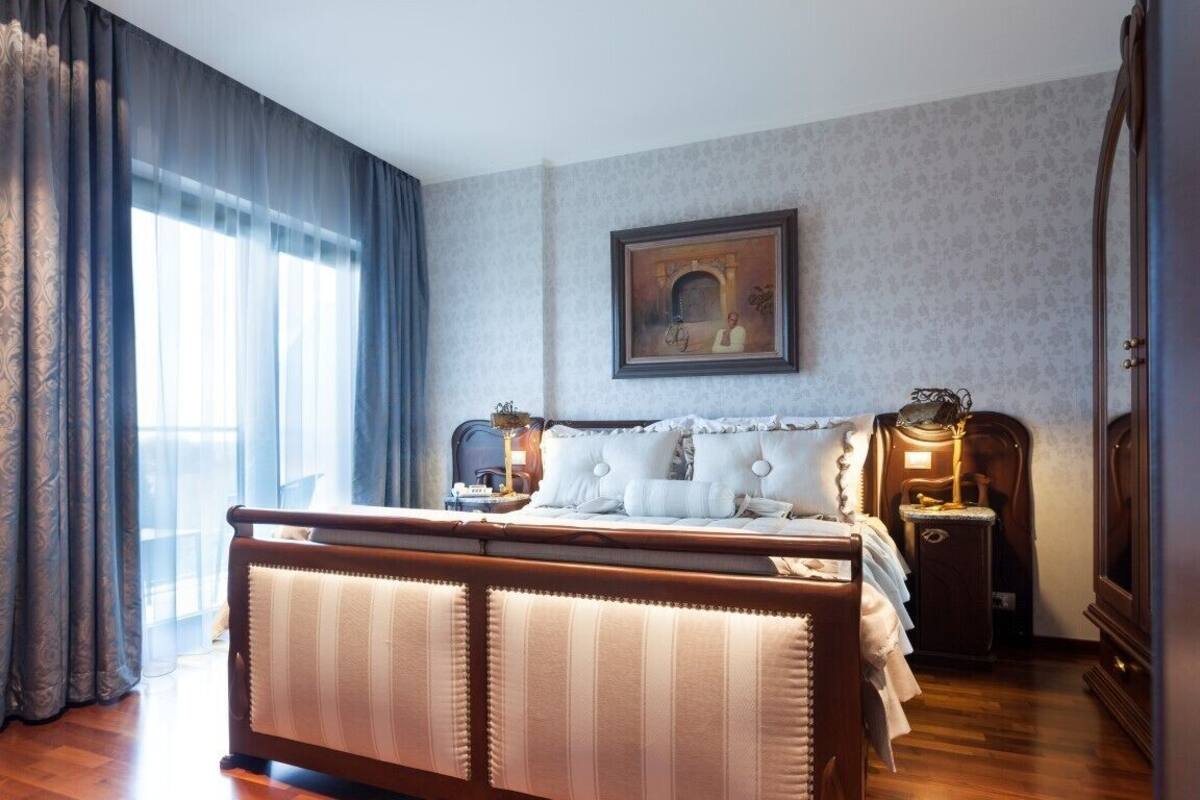Ein Schlafzimmer mit einem großen Bett, einem Nachttisch, einer Kommode, einem Wandbild und einem Fenster mit Vorhängen.