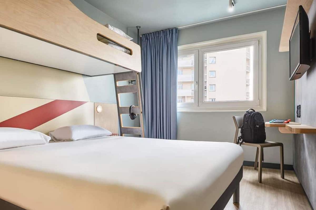 Habitación de hotel con literas, un escritorio, una silla, ventana con cortinas y televisor montado en la pared.