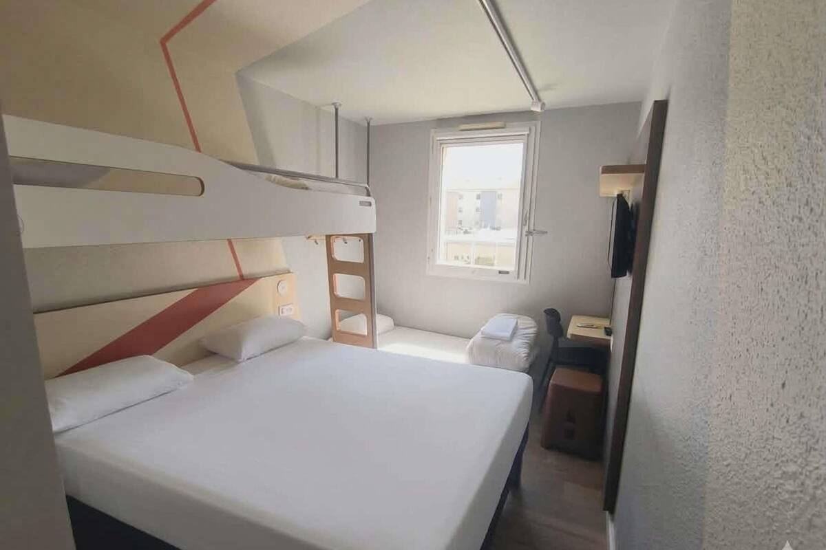 A compact hotel room with bunk beds, a desk, a chair, a small window, and a television.