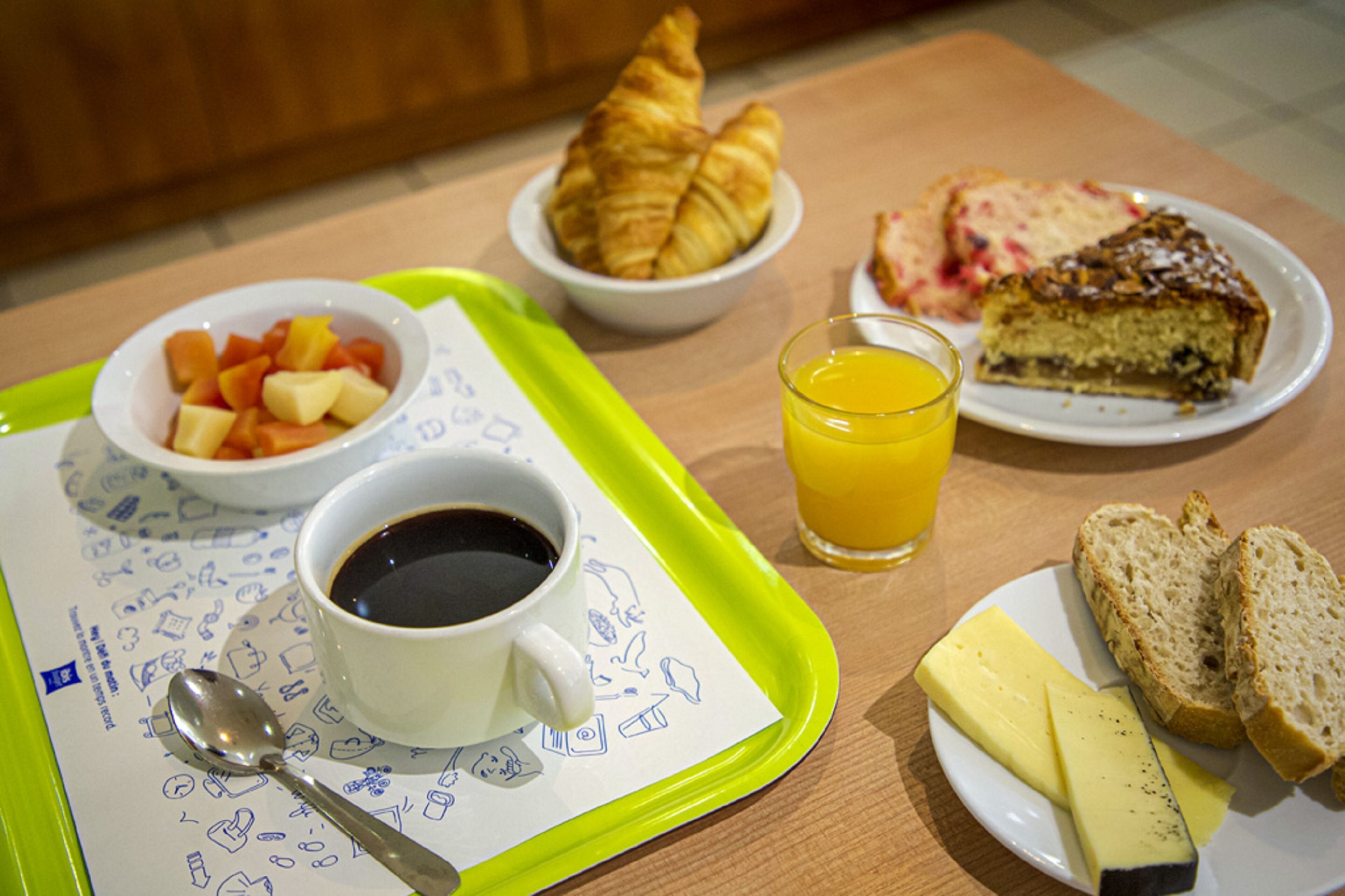 Bandeja de desayuno con croissants, fruta, queso, pan, un vaso de zumo de naranja y una taza de café sobre una mesa.