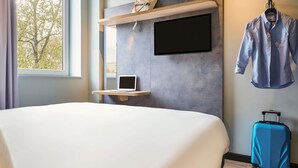 Una habitación de hotel con una cama, un escritorio con una computadora portátil, un televisor montado en la pared, un abrigo colgado y una maleta.