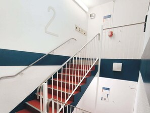 Una escalera con peldaños rojos y barandillas blancas, el número '2' en la pared y un detector de incendios a la derecha.